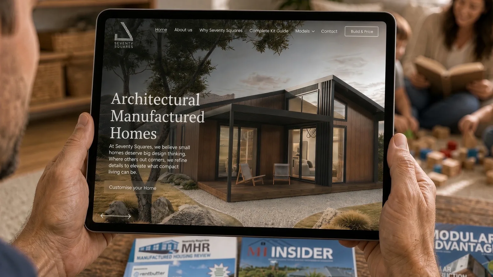 A buyer browsing a builder's website on a tablet at home, with US manufactured housing magazines on the table